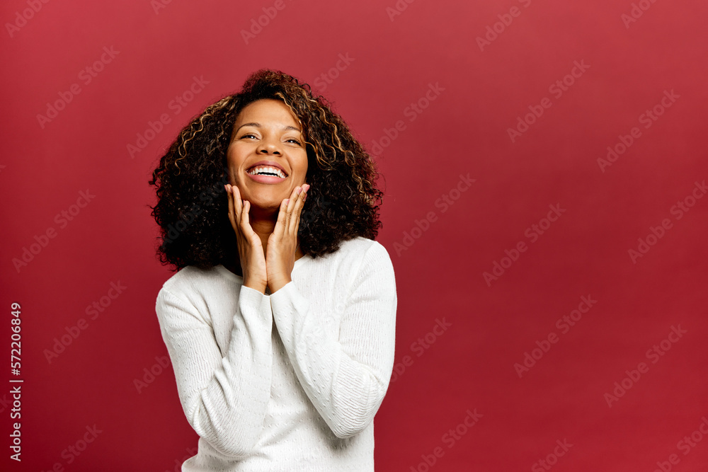 Smiling African woman touching cheeks with hands, posing on a red wall.