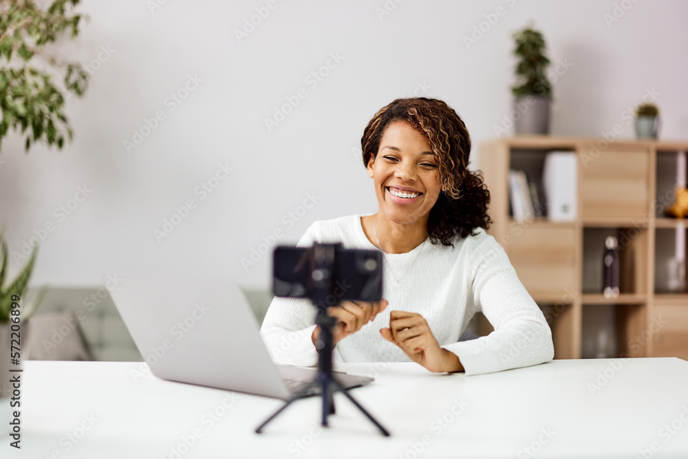 Smiling African American woman blogger recording video over the smartphone on a tripod at the workpl