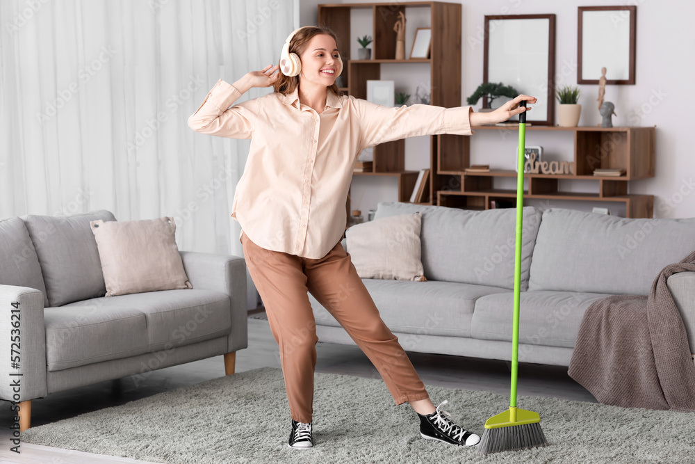 Young woman in headphones with broom dancing at home