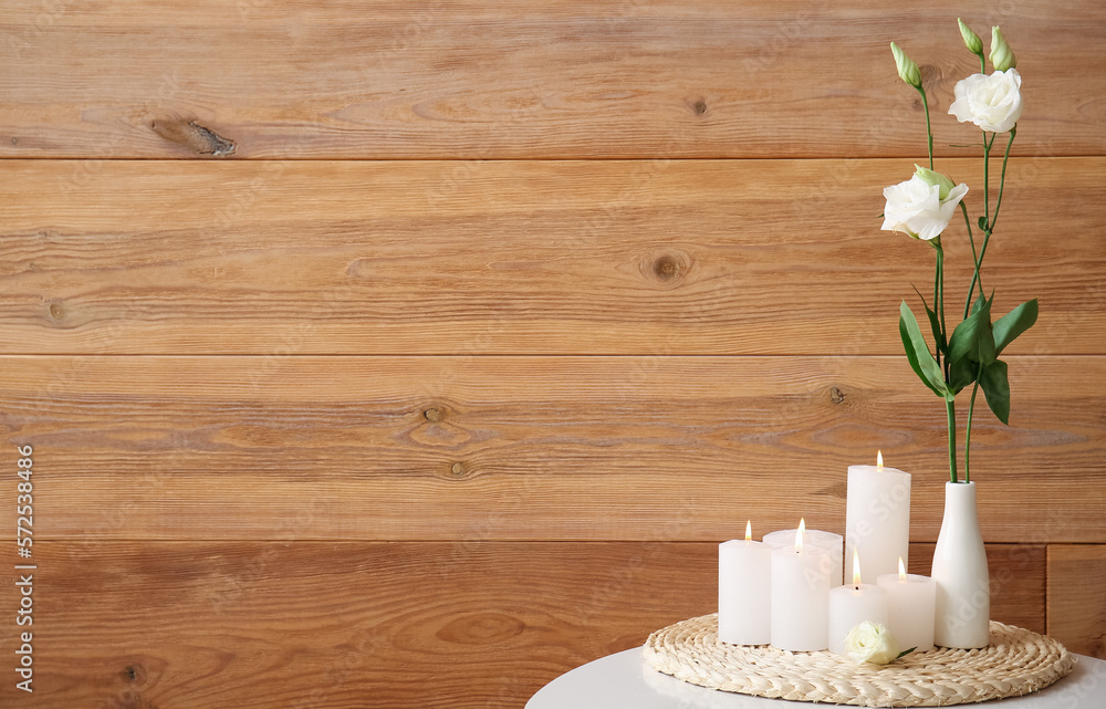 Burning candles and vase with flowers on table near wooden wall