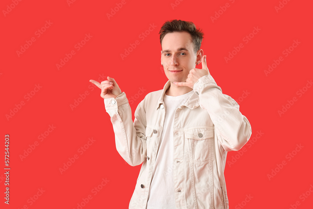 Cool young man showing  call me  gesture and pointing at something on red background