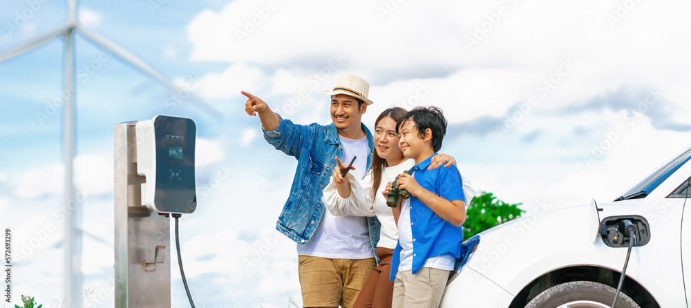 Concept of progressive happy family enjoying their time at wind farm with electric vehicle. Electric