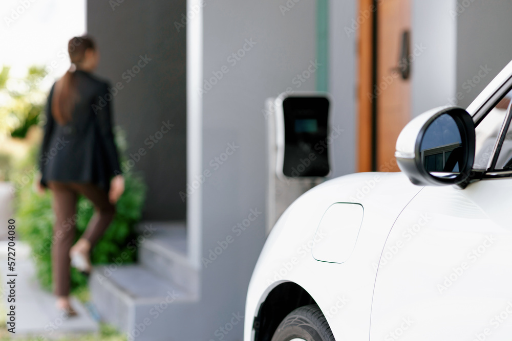 Focus image of electric vehicle recharging battery at home charging station with blurred woman walki