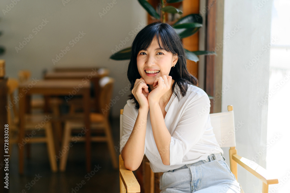 Brightly smiling beautiful young asian woman sitting in coffee cafe in sunny day