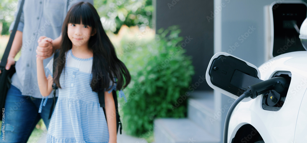 Focus electric car recharging at home charging station with blurred father and daughter walking in b