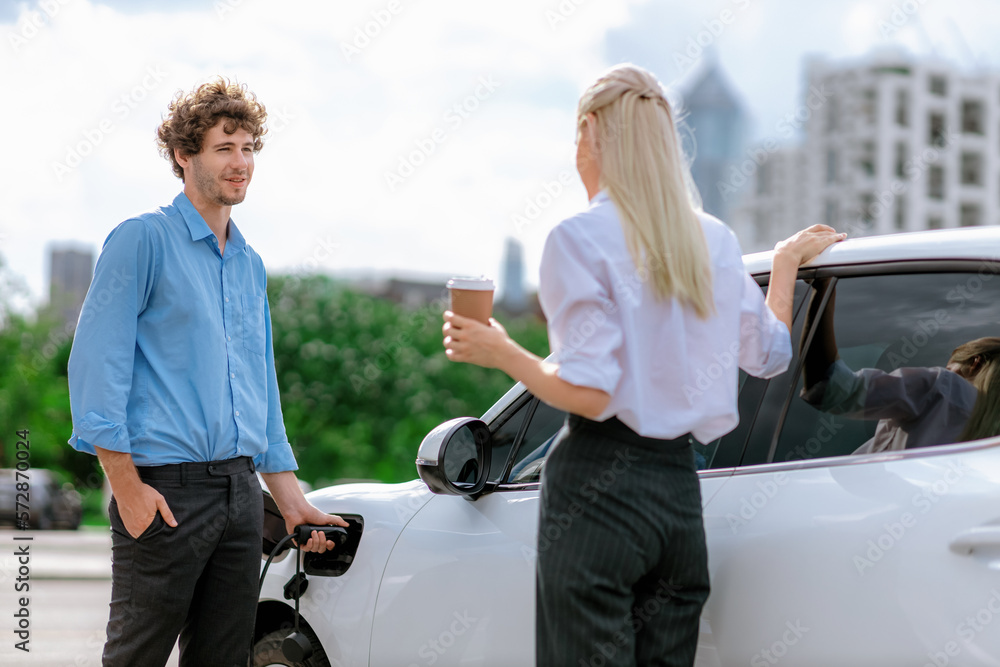 Progressive businessman and businesswoman with coffee, standing at electric car connected to chargin