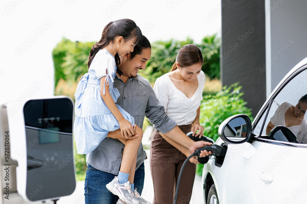 Progressive young parent teach daughter how to recharge or refuel EV car at home charging station. G