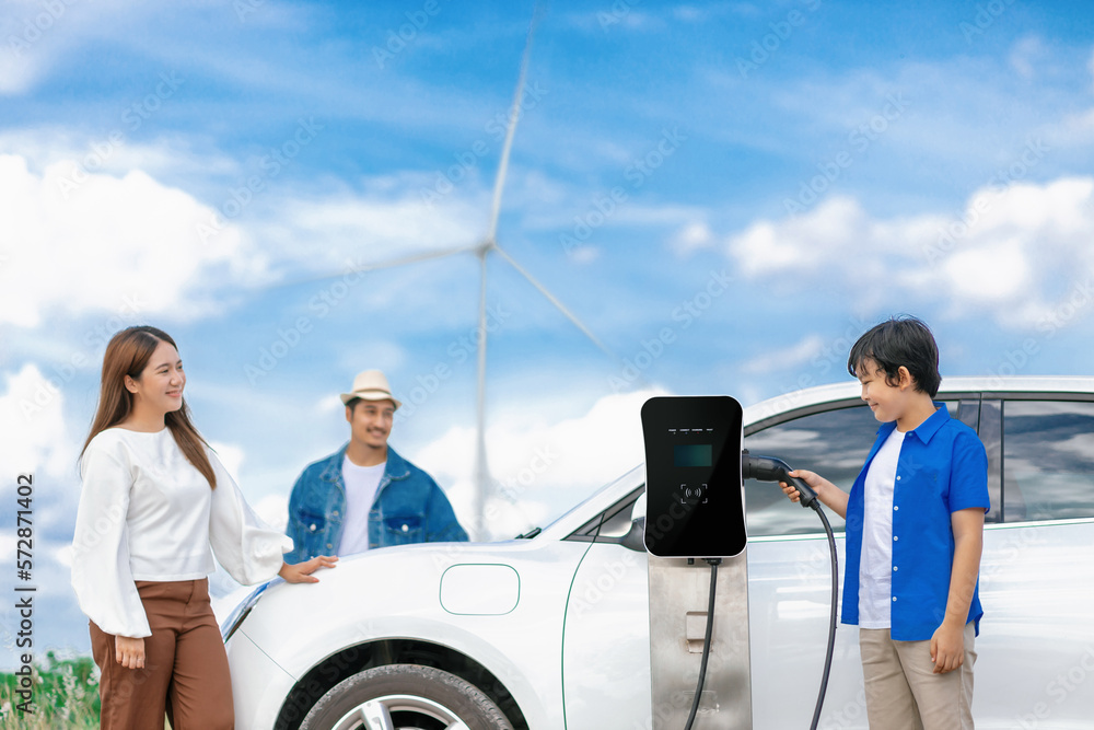 Concept of progressive happy family enjoying their time at wind farm with electric vehicle. Electric