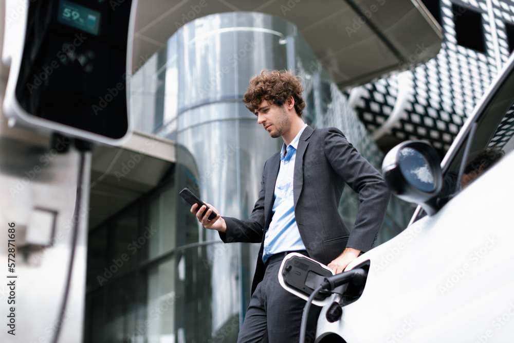 Below view of progressive businessman with electric car recharging at public charging station at mod