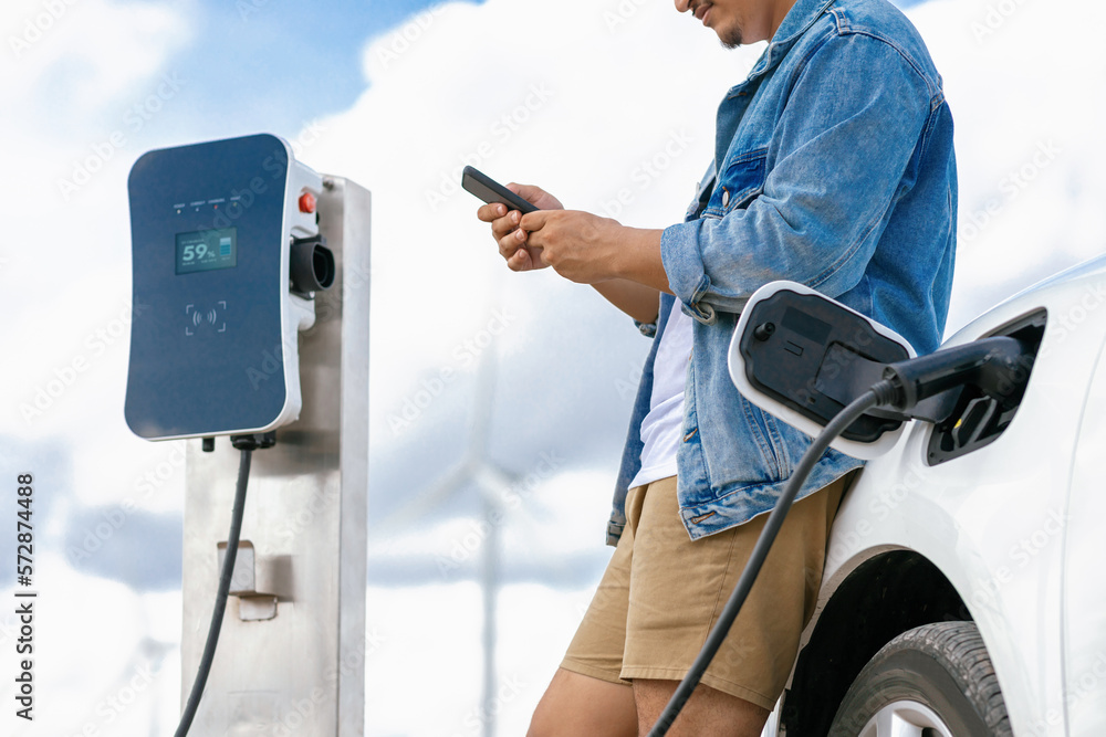 Progressive man with his electric car, EV car recharging energy from charging station on green field