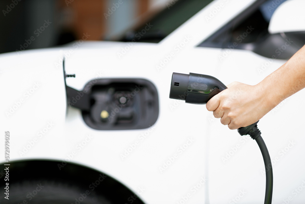 Focus hand holding EV charger plug with blurred background of progressive electric vehicle and socke