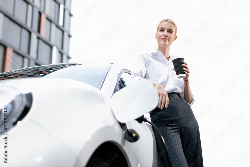 Businesswoman drinking coffee, leaning on electric vehicle recharging at public charging station wit