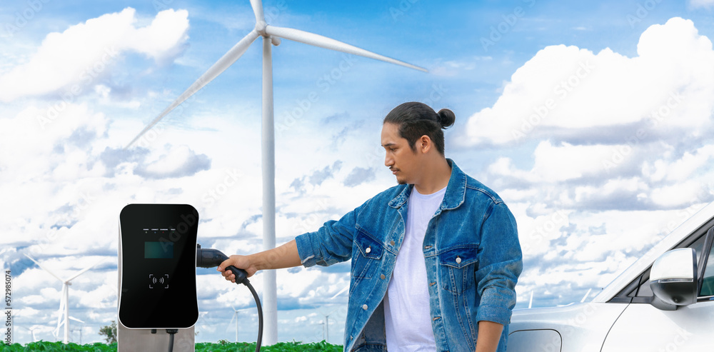 Progressive man with his electric car, EV car recharging energy from charging station on green field