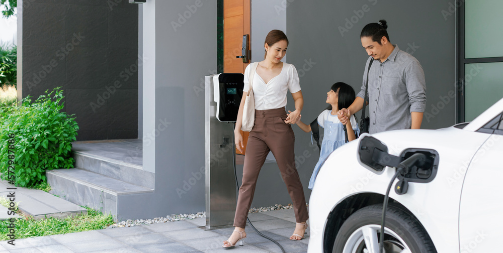 Progressive young parents and daughter with electric vehicle and home charging station. Green and cl