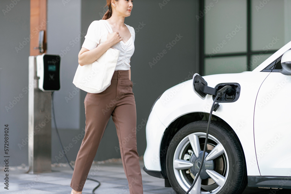 Progressive asian woman and electric car with home charging station. Concept of the use of electric 