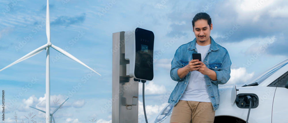 Progressive man with his electric car, EV car recharging energy from charging station on green field