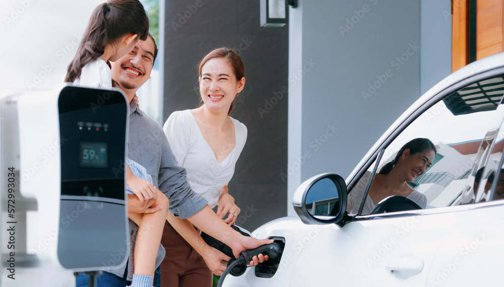 Progressive young parents and daughter with electric vehicle and home charging station. Green and cl