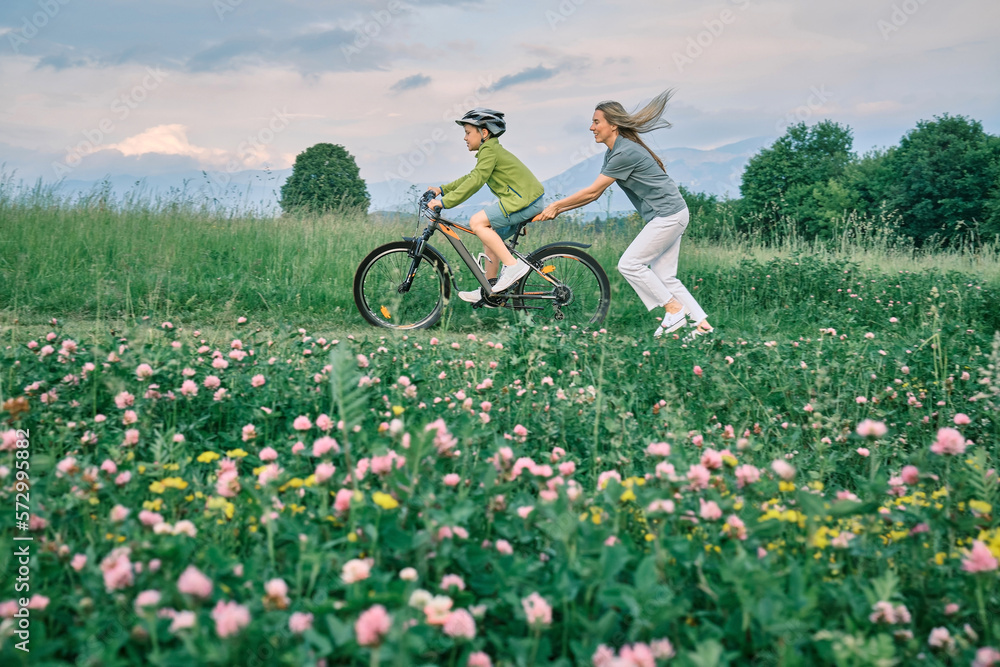 Mother teaching son to ride bicycle. Happy cute boy in helmet learn to riding a bike in park on gree