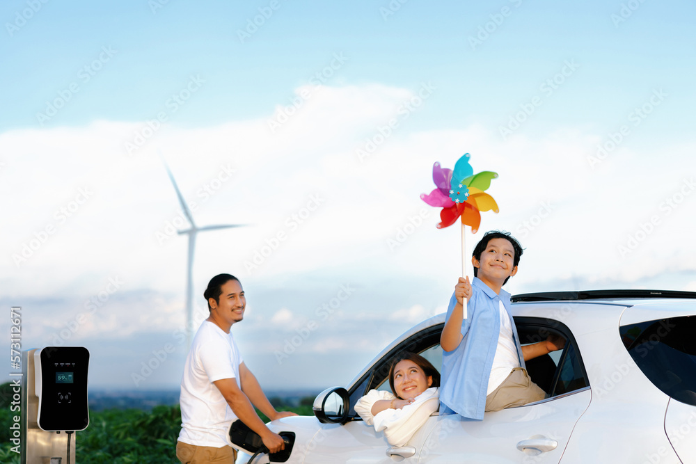 Concept of progressive happy family enjoying their time at wind farm with electric vehicle. Electric