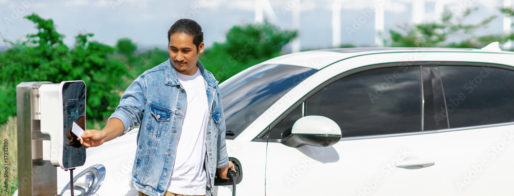 Progressive man with his electric car, EV car recharging energy from charging station on green field