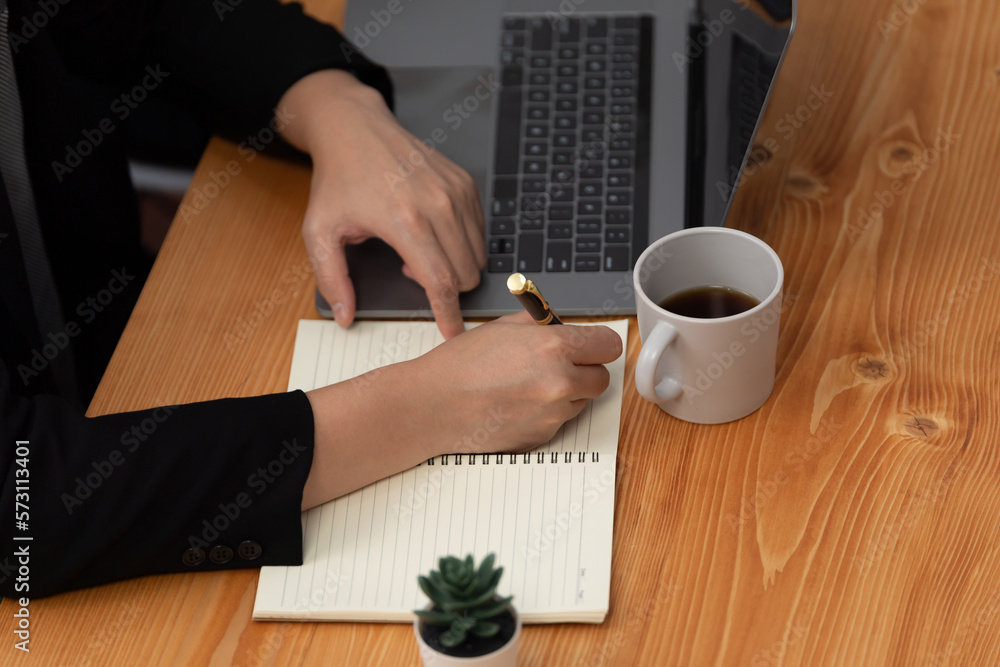 Closeup hand of secretary writing on notebook with laptop and coffee at her desk in harmony office. 