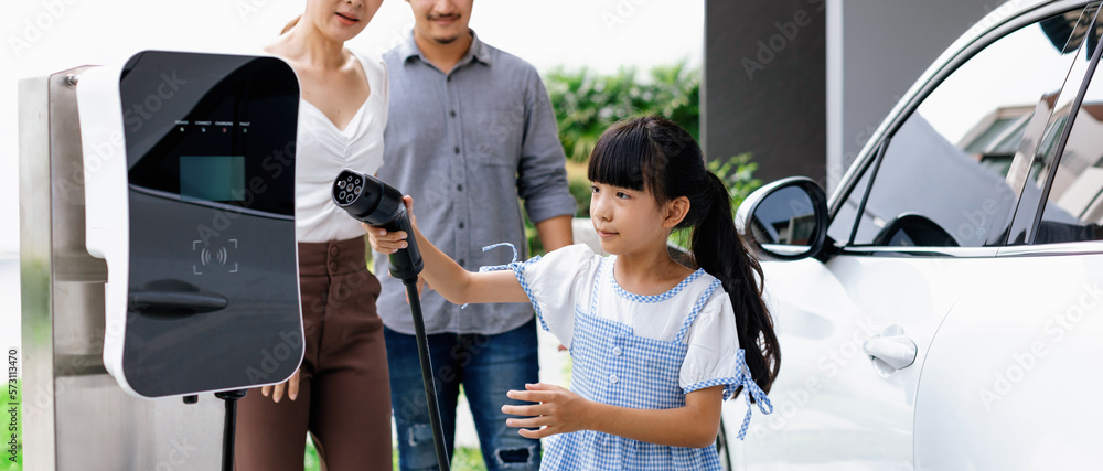 Progressive young parent teach daughter how to recharge or refuel EV car at home charging station. G