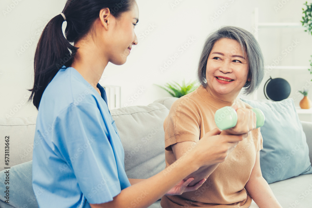 Caregiver helping contented senior woman exercise with dumbbell at home. Rehab for elder, senior med