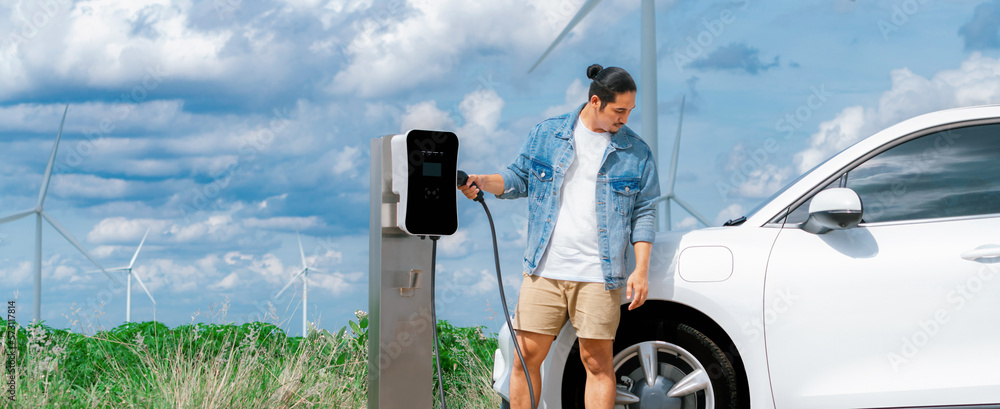 Progressive man with his electric car, EV car recharging energy from charging station on green field