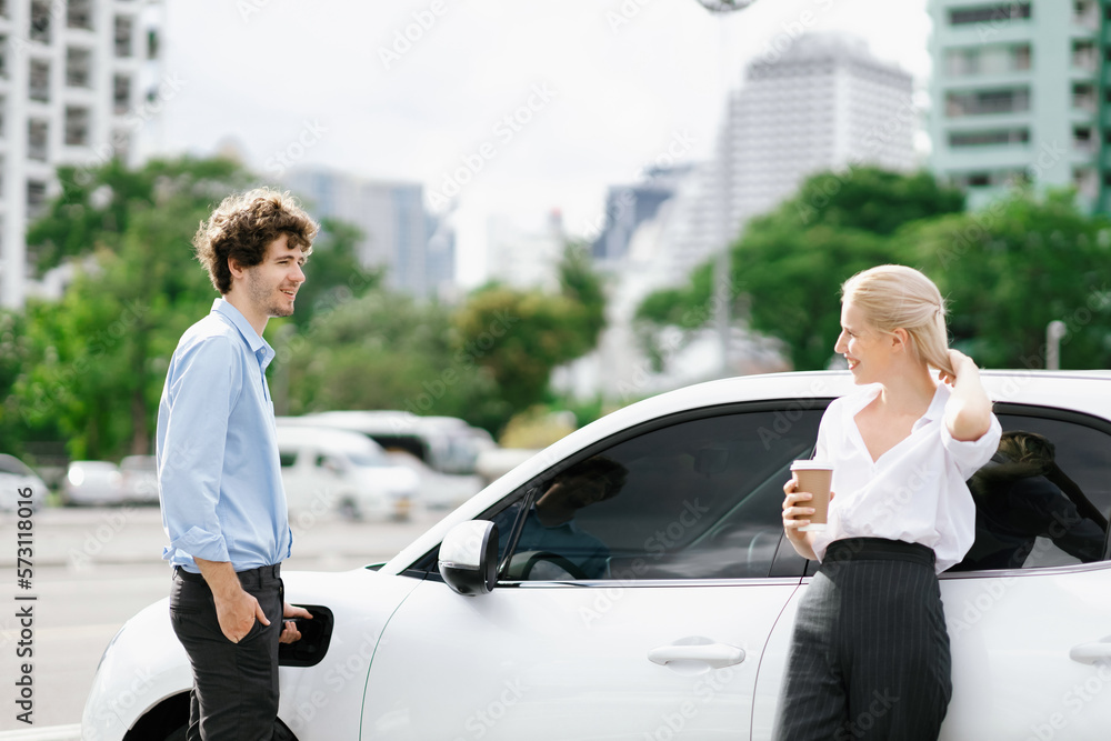 Progressive businessman and businesswoman with coffee, standing at electric car connected to chargin