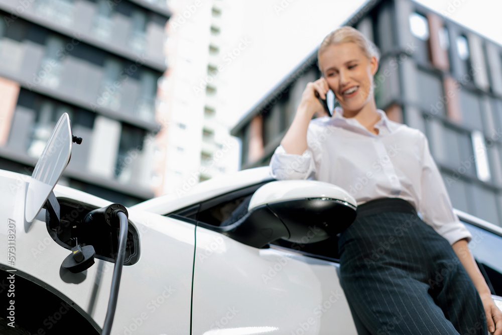 Focus charging-electric car with EV charger at charging station with blur businesswoman talking on p