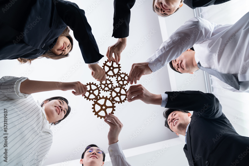 Hand holding wooden gear by businesspeople wearing suit for harmony synergy in office workplace. Bot