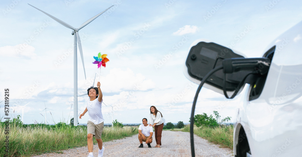 Concept of progressive happy family holding windmill toy and relax at wind farm with electric vehicl