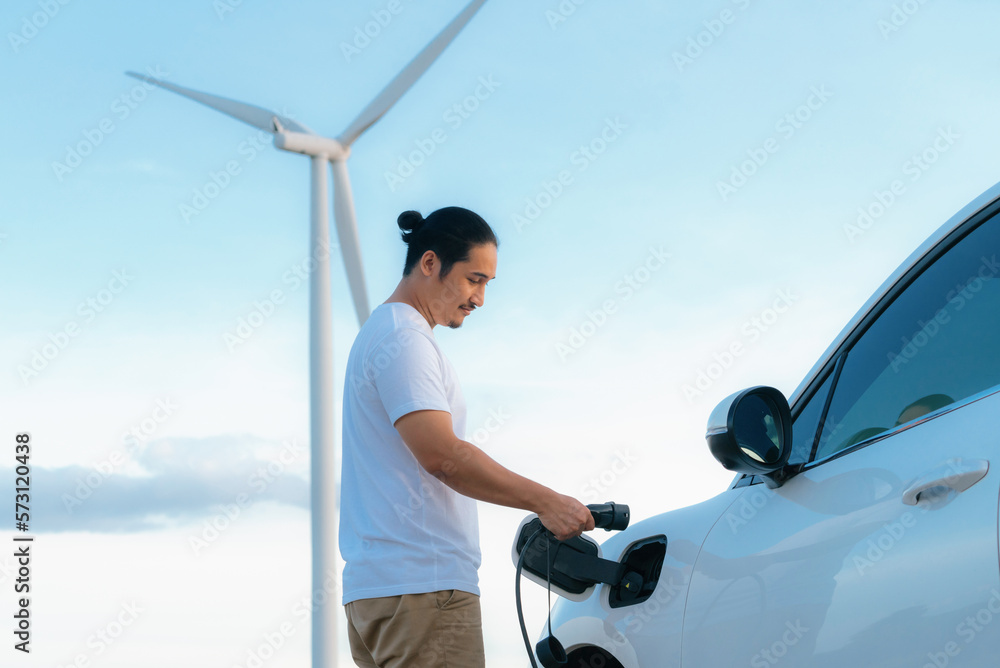 Progressive man with his electric car, EV car recharging energy from charging station on green field