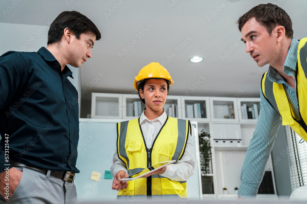 A team of competent engineers wearing safety equipment is working on blueprints with a tablet while 