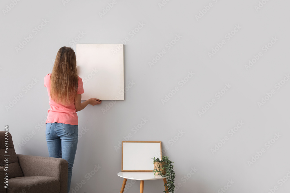 Young woman hanging blank poster on grey wall at home, back view