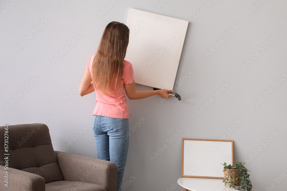 Young woman hanging blank poster on grey wall at home, back view