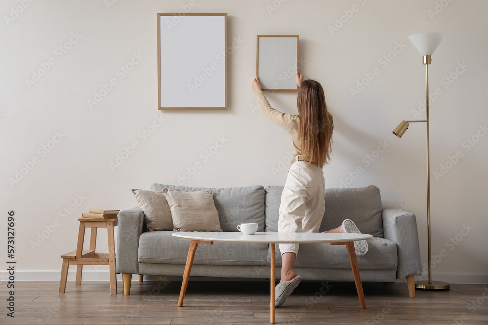 Young woman hanging blank frame on light wall at home, back view