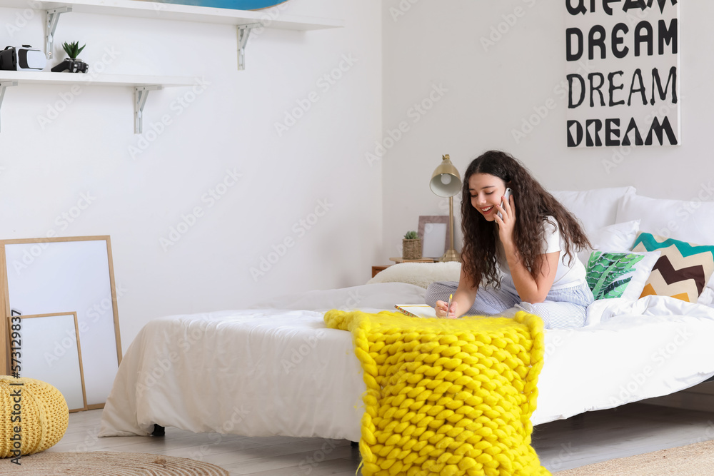 Teenage girl with notebook talking by mobile phone in bedroom