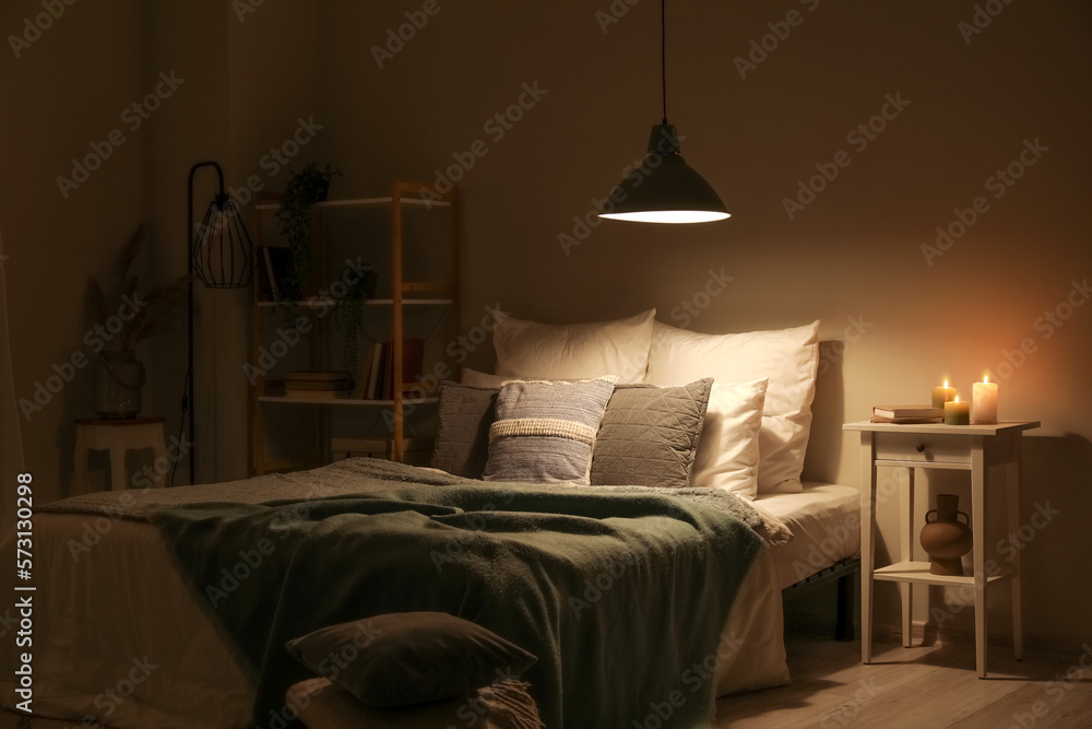 Interior of bedroom with green blankets on bed, burning candles and glowing lamp at night