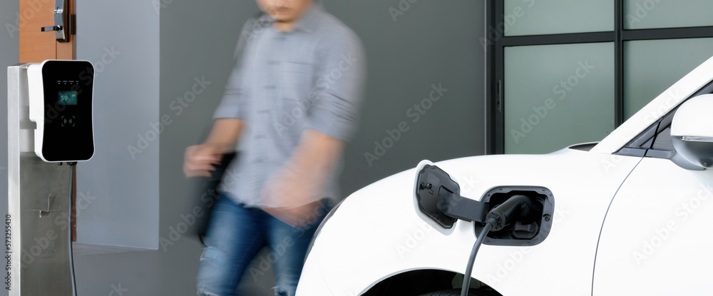 Focus electric car charging at home charging station with blurred progressive man walking in the bac