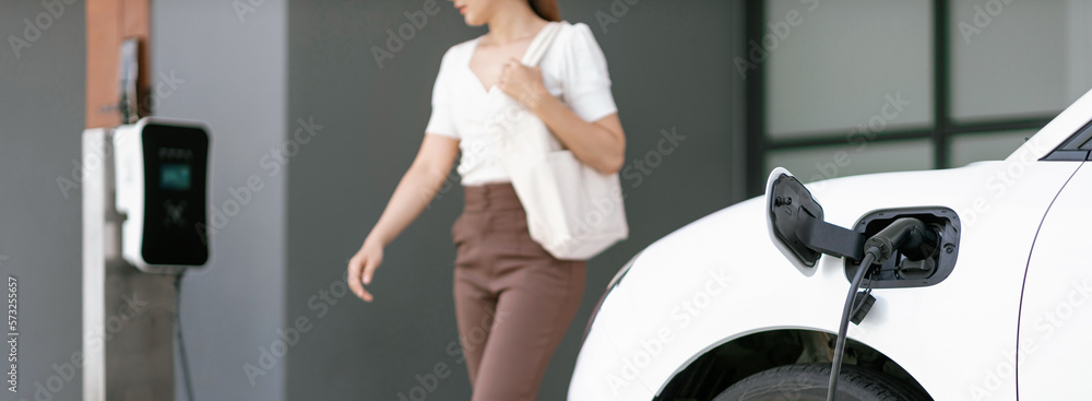 Focus image of electric vehicle recharging battery at home charging station with blurred woman walki