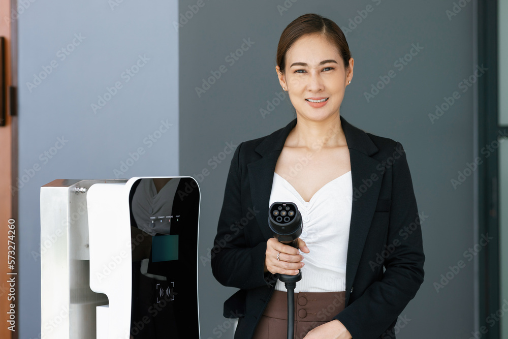 Asian woman holding and pointing an EV plug, a home charging station providing a sustainable power s