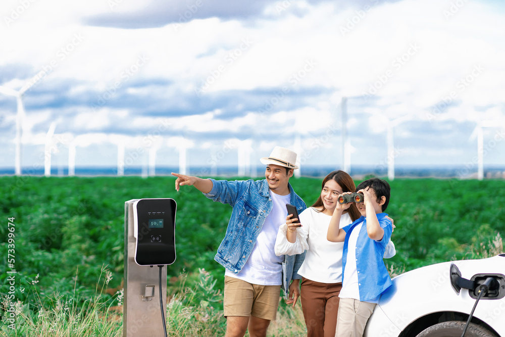 Concept of progressive happy family enjoying their time at wind farm with electric vehicle. Electric