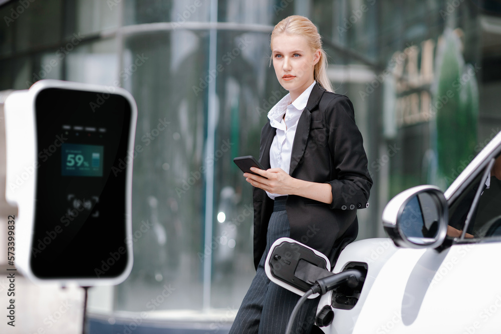 Businesswoman wearing black suit using smartphone, leaning on electric car recharge battery at charg