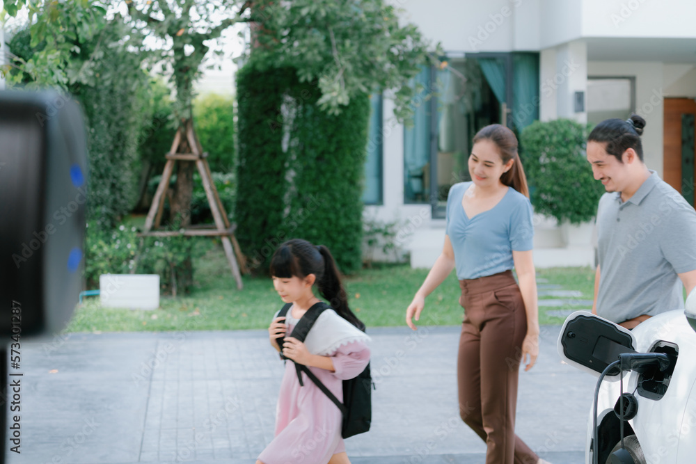 Progressive young parents and daughter with electric vehicle and home charging station. Green and cl