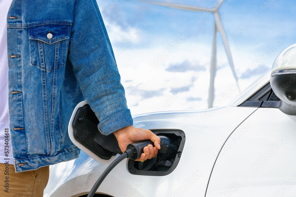 Progressive man with his electric car, EV car recharging energy from charging station on green field