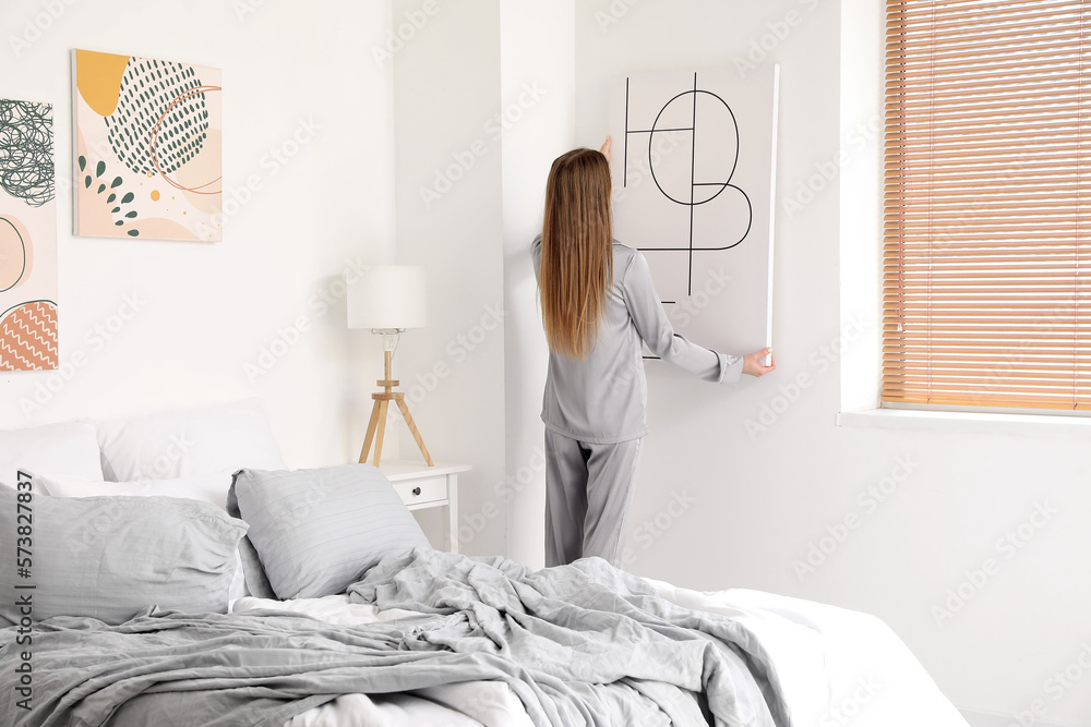 Young woman hanging poster on light wall in bedroom