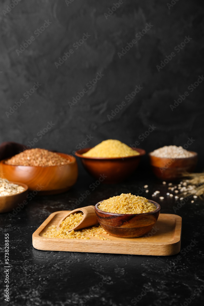 Wooden board with bowl and scoop of bulgur on dark background