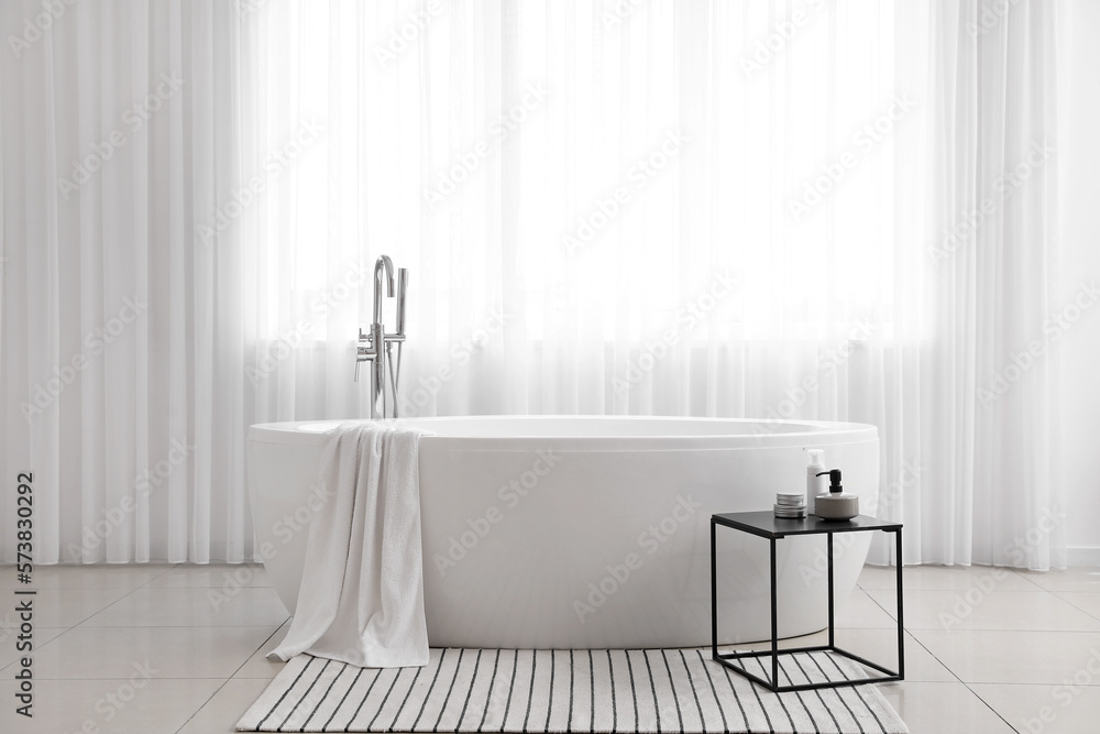 Bathtub and table with cosmetic products in light bathroom