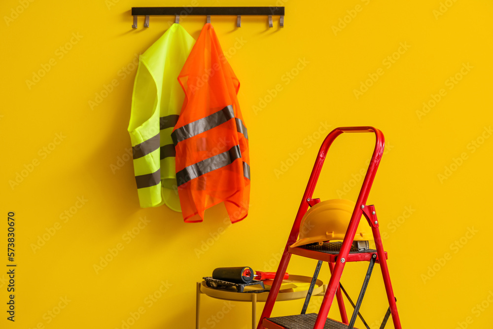 Reflective vests, stepladder and table with builders tools near yellow wall